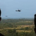 Two military attack helicopters, consistent with AH-64 Apache design, are flying over a dense forested landscape under overcast skies. Two silhouetted observers in the foreground watch the helicopters from an elevated vantage point. The scene appears to depict a military training exercise or demonstration.