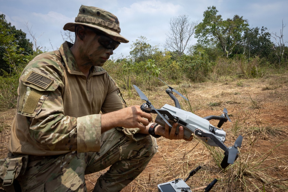 A U.S. military service member in MultiCam/OCP camouflage uniform is preparing or inspecting a small commercial quadcopter drone, likely a DJI Mavic series, in an outdoor field environment. The soldier has face camouflage paint applied and is wearing a boonie hat and tactical sunglasses. A drone controller/tablet is visible on the ground nearby.
