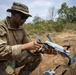 A U.S. military service member in MultiCam/OCP camouflage uniform is preparing or inspecting a small commercial quadcopter drone, likely a DJI Mavic series, in an outdoor field environment. The soldier has face camouflage paint applied and is wearing a boonie hat and tactical sunglasses. A drone controller/tablet is visible on the ground nearby.