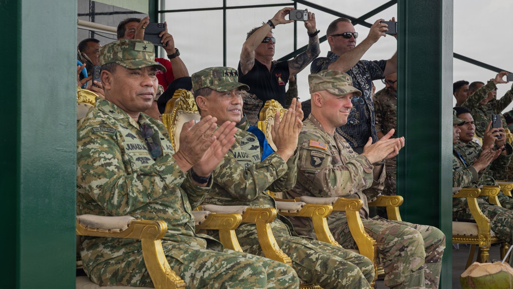 A group of military officers from at least two different nations are seated in ornate gold chairs at what appears to be an official military ceremony or demonstration. Three senior officers in the foreground are applauding, with two wearing camouflage uniforms bearing star insignia and one wearing a US Army uniform with a 101st Airborne Division patch. Civilians and additional military personnel in the background are photographing the event with cameras and smartphones.