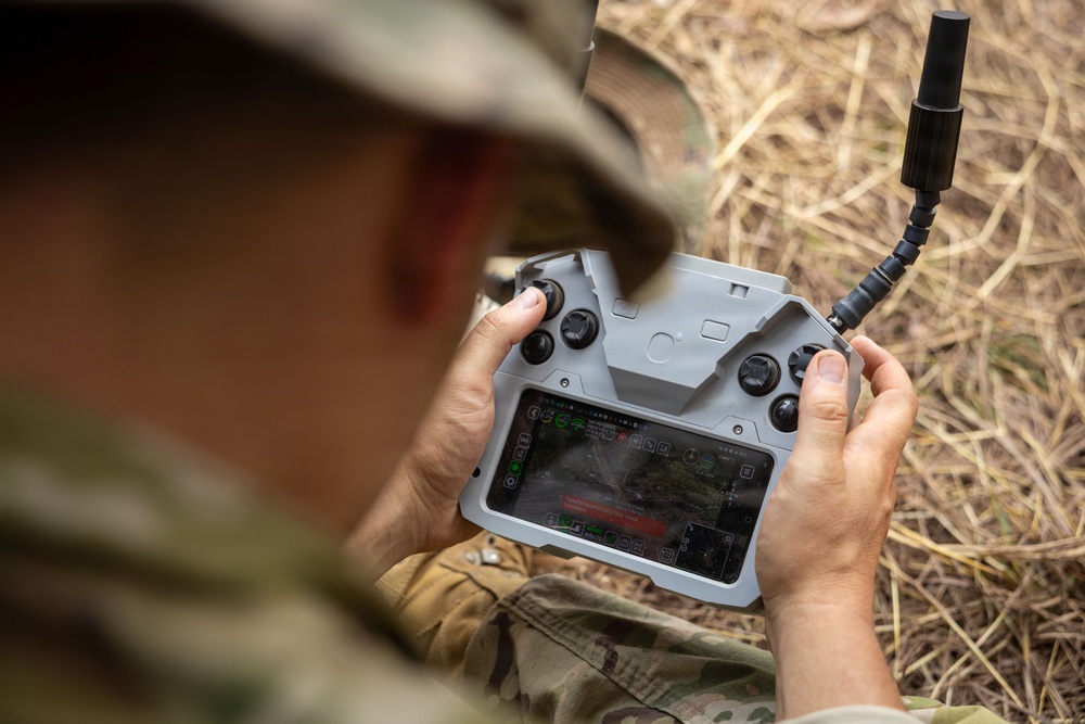 A person wearing military camouflage uniform and hat is operating a drone remote controller with an integrated touchscreen display. The controller appears to be a DJI-style unit with dual joysticks, buttons, and a built-in screen showing a live feed or navigation interface. The setting appears to be an outdoor field environment with dry grass visible in the background.