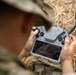 A person wearing military camouflage uniform and hat is operating a drone remote controller with an integrated touchscreen display. The controller appears to be a DJI-style unit with dual joysticks, buttons, and a built-in screen showing a live feed or navigation interface. The setting appears to be an outdoor field environment with dry grass visible in the background.
