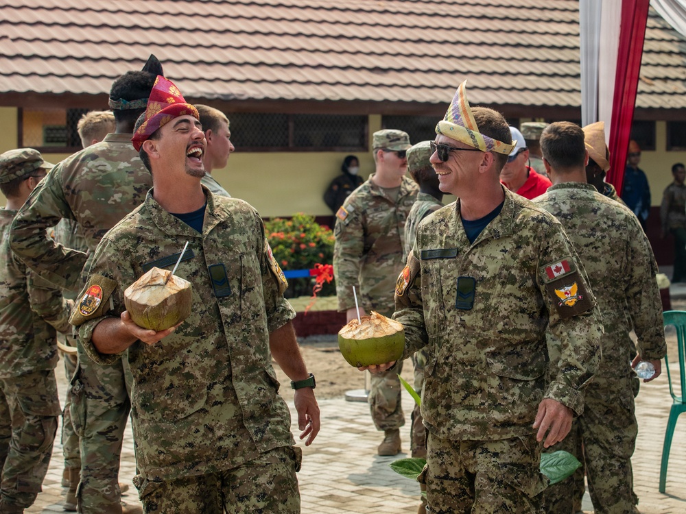 Two military personnel in digital camouflage uniforms are laughing and holding fresh coconuts with straws, wearing traditional decorative headwear. One individual has a visible Canadian flag patch on their sleeve, suggesting a multinational military exercise or cultural exchange event. The setting appears to be an outdoor courtyard of a building, with additional military personnel visible in the background.
