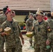 Two military personnel in digital camouflage uniforms are laughing and holding fresh coconuts with straws, wearing traditional decorative headwear. One individual has a visible Canadian flag patch on their sleeve, suggesting a multinational military exercise or cultural exchange event. The setting appears to be an outdoor courtyard of a building, with additional military personnel visible in the background.