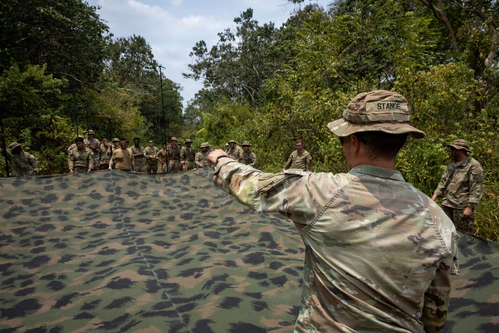A military instructor identified by the name tape 'STANGE' on his hat is directing a group of soldiers during what appears to be a field training exercise in a jungle or forested environment. The central figure is holding or spreading a large camouflage tarpaulin/net while gesturing toward a group of approximately 15-20 soldiers standing in the background. The setting suggests a tropical or subtropical region based on the dense vegetation.