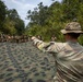A military instructor identified by the name tape 'STANGE' on his hat is directing a group of soldiers during what appears to be a field training exercise in a jungle or forested environment. The central figure is holding or spreading a large camouflage tarpaulin/net while gesturing toward a group of approximately 15-20 soldiers standing in the background. The setting suggests a tropical or subtropical region based on the dense vegetation.