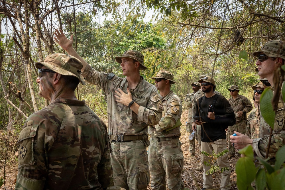 A group of U.S. Army soldiers in MultiCam/OCP camouflage uniforms are gathered in a wooded, dry-season environment. A central soldier wearing a boonie hat is gesturing with his raised hand, appearing to brief or instruct the group. One civilian or non-uniformed individual in a black shirt is also present among the group.