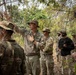 A group of U.S. Army soldiers in MultiCam/OCP camouflage uniforms are gathered in a wooded, dry-season environment. A central soldier wearing a boonie hat is gesturing with his raised hand, appearing to brief or instruct the group. One civilian or non-uniformed individual in a black shirt is also present among the group.
