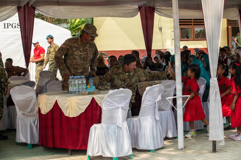 The image shows a military community engagement event, likely in Southeast Asia, where U.S. Army soldiers in camouflage uniforms are interacting with local civilians under a decorated tent canopy. The setting appears to be a school or community facility, with children in red uniforms and women in hijabs gathered as attendees. The event has a formal ceremonial setup with decorated tables and chairs.