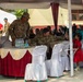 The image shows a military community engagement event, likely in Southeast Asia, where U.S. Army soldiers in camouflage uniforms are interacting with local civilians under a decorated tent canopy. The setting appears to be a school or community facility, with children in red uniforms and women in hijabs gathered as attendees. The event has a formal ceremonial setup with decorated tables and chairs.