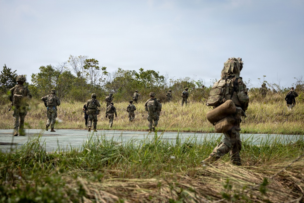 A group of military personnel in full combat gear and camouflage uniforms are moving across a paved surface toward a treeline in what appears to be a tropical or subtropical environment. The soldiers are carrying heavy rucksacks and tactical equipment, suggesting a field training exercise or operational movement. The scene is photographed from a low angle with a shallow depth of field, emphasizing the foreground soldier.