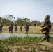 A group of military personnel in full combat gear and camouflage uniforms are moving across a paved surface toward a treeline in what appears to be a tropical or subtropical environment. The soldiers are carrying heavy rucksacks and tactical equipment, suggesting a field training exercise or operational movement. The scene is photographed from a low angle with a shallow depth of field, emphasizing the foreground soldier.