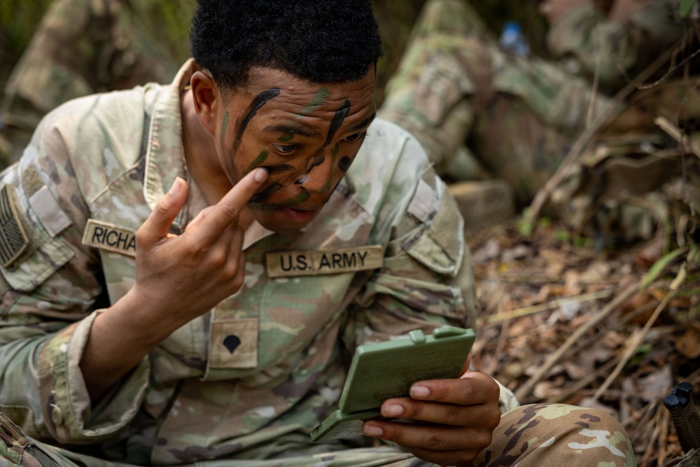 A U.S. Army soldier is applying camouflage face paint while using a small military-issue mirror for guidance. The soldier, whose name tape reads 'RICHA-' (partially visible), is in a woodland/outdoor training environment. Green, black, and brown camouflage paint has already been partially applied to the face.