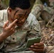 A U.S. Army soldier is applying camouflage face paint while using a small military-issue mirror for guidance. The soldier, whose name tape reads 'RICHA-' (partially visible), is in a woodland/outdoor training environment. Green, black, and brown camouflage paint has already been partially applied to the face.