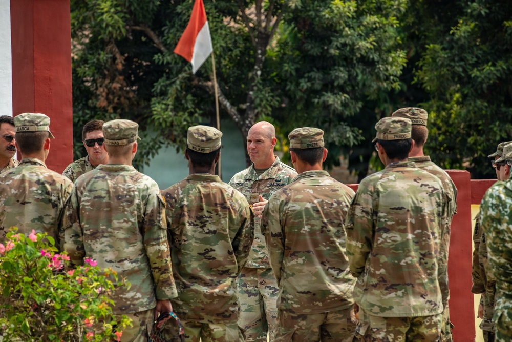 A group of U.S. Army soldiers in MultiCam/OCP camouflage uniforms are gathered outdoors, appearing to receive a briefing or address from a senior soldier facing them. A red and white flag — consistent with the Indonesian flag — is visible in the background, suggesting this may be a joint military exercise or visit in Indonesia. The setting includes tropical vegetation and a red-painted structure.