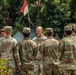 A group of U.S. Army soldiers in MultiCam/OCP camouflage uniforms are gathered outdoors, appearing to receive a briefing or address from a senior soldier facing them. A red and white flag — consistent with the Indonesian flag — is visible in the background, suggesting this may be a joint military exercise or visit in Indonesia. The setting includes tropical vegetation and a red-painted structure.
