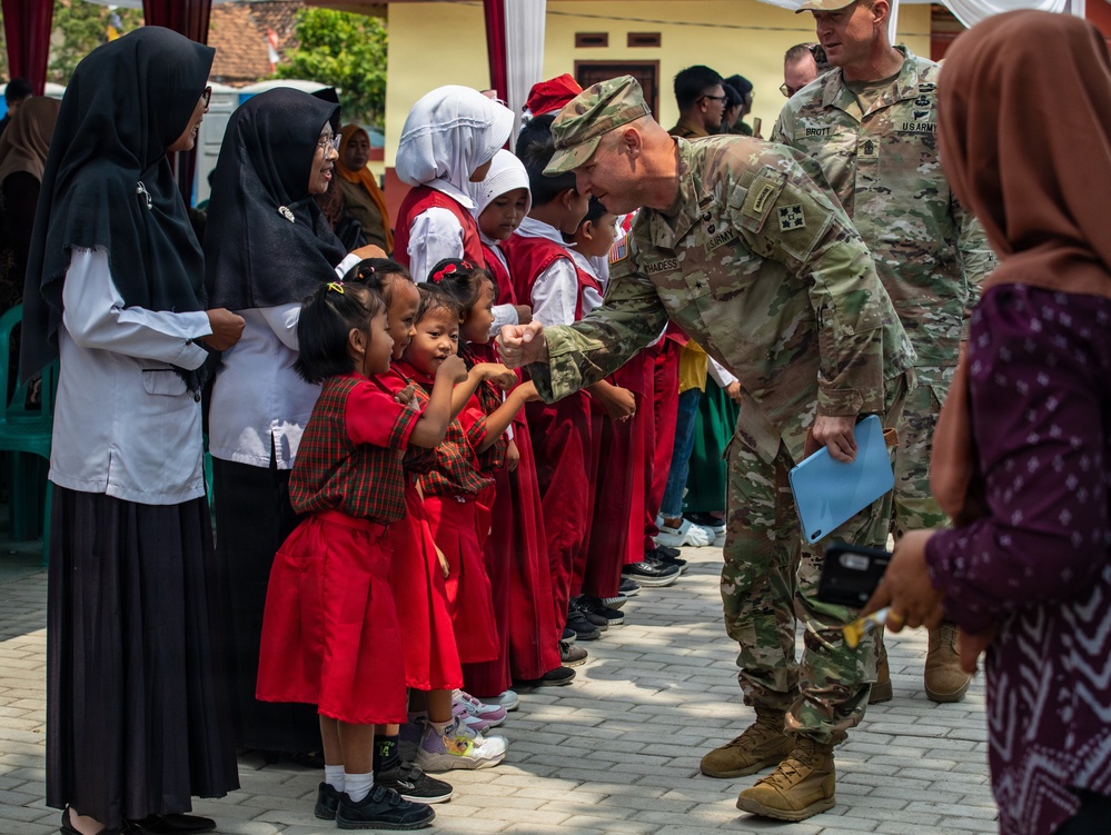 A U.S. Army soldier in MultiCam camouflage uniform is fist-bumping young children dressed in red school uniforms during what appears to be a community engagement or humanitarian visit. A second U.S. Army soldier with the name tape 'BROTT' is visible in the background. The setting appears to be a school or community gathering in Southeast Asia, likely Indonesia, based on the hijab-wearing women and local architectural style.