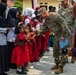 A U.S. Army soldier in MultiCam camouflage uniform is fist-bumping young children dressed in red school uniforms during what appears to be a community engagement or humanitarian visit. A second U.S. Army soldier with the name tape 'BROTT' is visible in the background. The setting appears to be a school or community gathering in Southeast Asia, likely Indonesia, based on the hijab-wearing women and local architectural style.