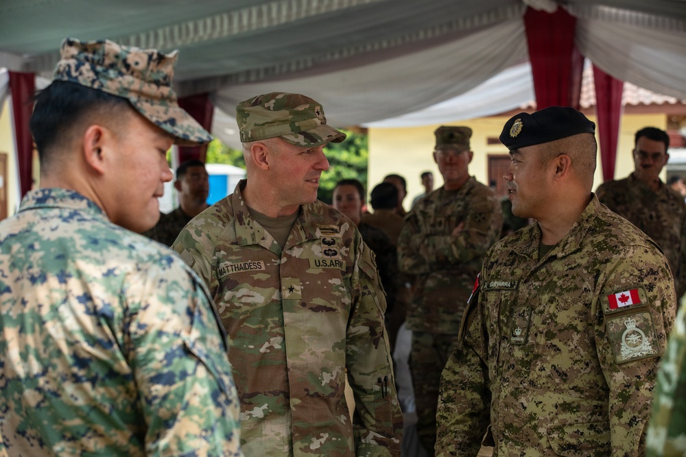 A multinational military gathering showing personnel from at least three nations in conversation under a tent or canopy structure. A U.S. Army officer identified by name tape as 'MATTHAIDESS' with a rank insignia is engaged in discussion with a Canadian Armed Forces soldier wearing a black beret and Canadian flag patch. A third soldier in digital camouflage pattern is visible on the left side of the frame.