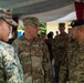 A multinational military gathering showing personnel from at least three nations in conversation under a tent or canopy structure. A U.S. Army officer identified by name tape as 'MATTHAIDESS' with a rank insignia is engaged in discussion with a Canadian Armed Forces soldier wearing a black beret and Canadian flag patch. A third soldier in digital camouflage pattern is visible on the left side of the frame.