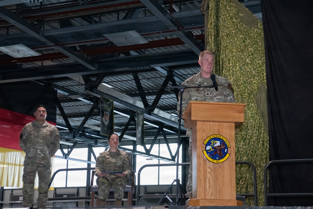 A military ceremony is taking place inside what appears to be a hangar or large indoor facility. A uniformed service member is speaking at a wooden podium bearing the insignia of the 721st Aerial Port Squadron (721 APS). Two other uniformed personnel stand and sit on a stage behind the speaker, with a German flag visible in the background.