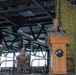 A military ceremony is taking place inside what appears to be a hangar or large indoor facility. A uniformed service member is speaking at a wooden podium bearing the insignia of the 721st Aerial Port Squadron (721 APS). Two other uniformed personnel stand and sit on a stage behind the speaker, with a German flag visible in the background.
