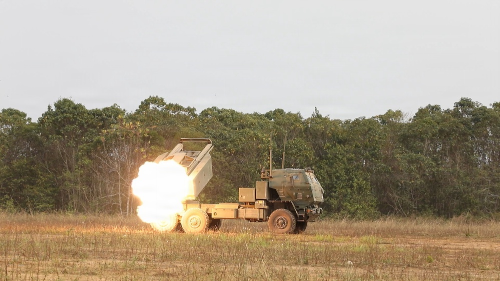 A multiple launch rocket system (MLRS), consistent with the M142 HIMARS configuration, is captured mid-fire on an open grassy field. A large fireball and exhaust plume are visible at the launcher pod as a rocket is being fired. The vehicle is positioned against a backdrop of dense tropical or subtropical tree line under an overcast sky.