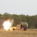 A multiple launch rocket system (MLRS), consistent with the M142 HIMARS configuration, is captured mid-fire on an open grassy field. A large fireball and exhaust plume are visible at the launcher pod as a rocket is being fired. The vehicle is positioned against a backdrop of dense tropical or subtropical tree line under an overcast sky.