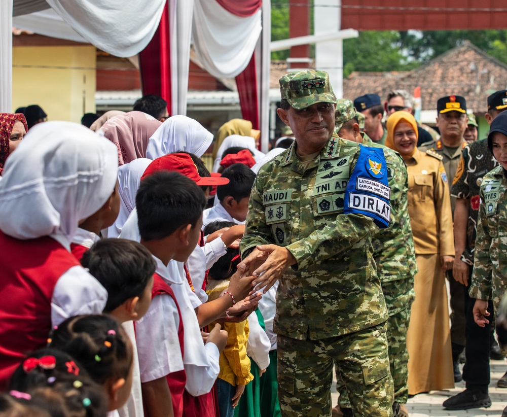 A senior Indonesian Army (TNI AD) officer named 'NAUDI' wearing the 'DIRLAT' designation patch and 'LATGABMA SUPER GARUDA SHIELD 2025' insignia greets schoolchildren at what appears to be a military-civic community event. The officer is surrounded by children in school uniforms and accompanied by other military and civilian officials. The setting appears to be an outdoor ceremonial area decorated with red and white Indonesian national colors.