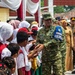 A senior Indonesian Army (TNI AD) officer named 'NAUDI' wearing the 'DIRLAT' designation patch and 'LATGABMA SUPER GARUDA SHIELD 2025' insignia greets schoolchildren at what appears to be a military-civic community event. The officer is surrounded by children in school uniforms and accompanied by other military and civilian officials. The setting appears to be an outdoor ceremonial area decorated with red and white Indonesian national colors.