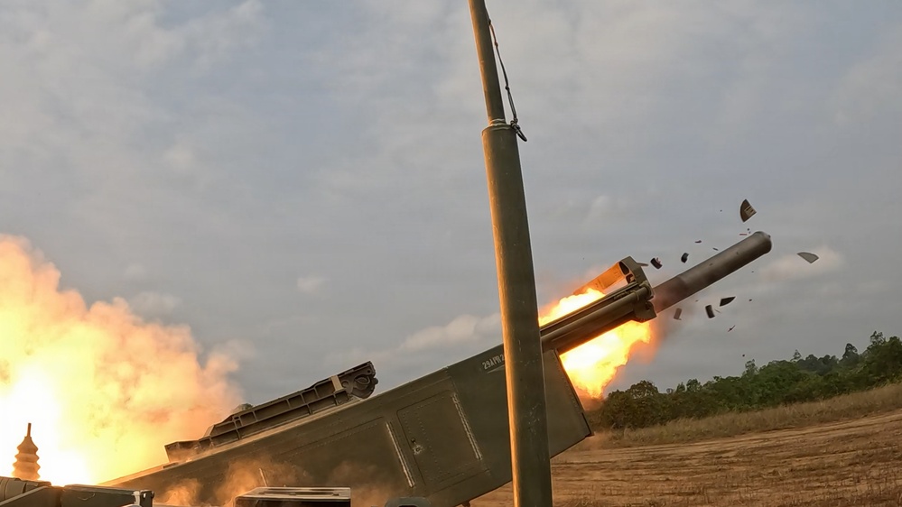 The image captures a multiple launch rocket system (MLRS) firing a rocket, with a large fireball and debris visible at the launch tube exit. The vehicle appears to be a military rocket artillery platform photographed at close range during an active launch. The scene takes place in an open field or training area with treeline visible in the background.
