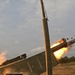 The image captures a multiple launch rocket system (MLRS) firing a rocket, with a large fireball and debris visible at the launch tube exit. The vehicle appears to be a military rocket artillery platform photographed at close range during an active launch. The scene takes place in an open field or training area with treeline visible in the background.