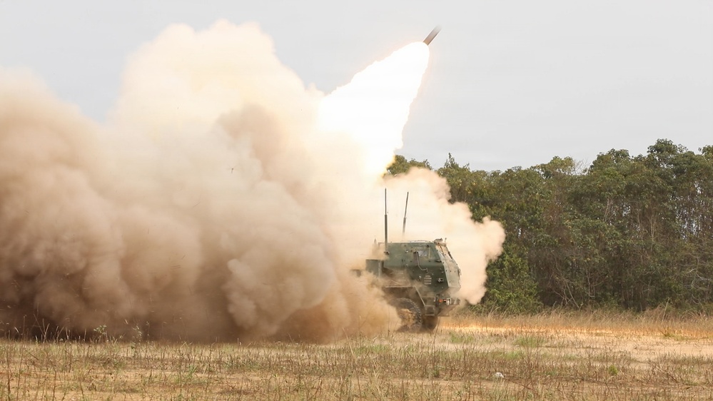 A multiple launch rocket system (MLRS) is captured mid-fire, with a rocket visibly ascending into the overcast sky. A large plume of tan/beige smoke and dust billows around the tracked military vehicle, which is painted in olive drab camouflage. The setting appears to be an open field or firing range with sparse dry grass and tree line in the background.