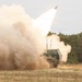 A multiple launch rocket system (MLRS) is captured mid-fire, with a rocket visibly ascending into the overcast sky. A large plume of tan/beige smoke and dust billows around the tracked military vehicle, which is painted in olive drab camouflage. The setting appears to be an open field or firing range with sparse dry grass and tree line in the background.
