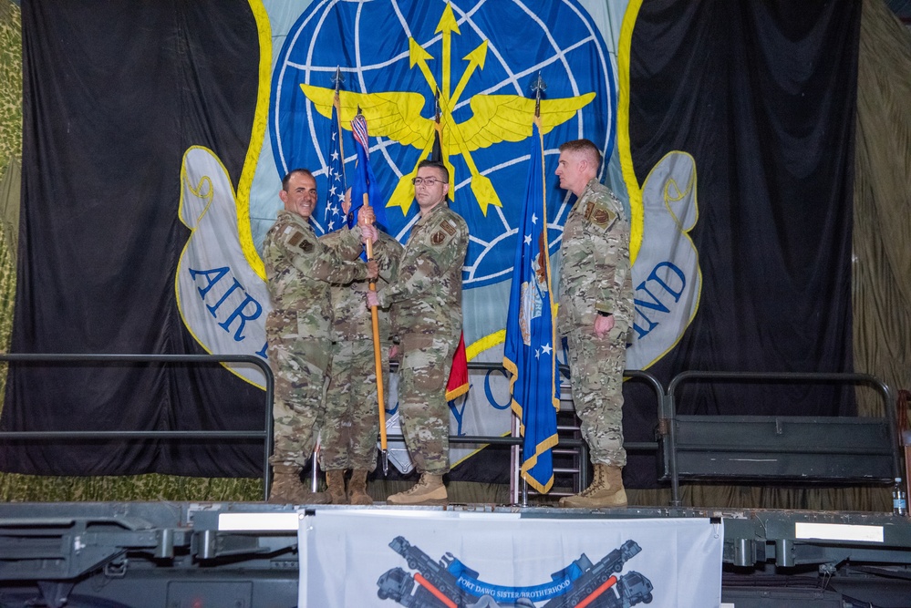 Three military personnel in camouflage uniforms are participating in what appears to be a change of command or guidon ceremony on a stage. A large unit emblem banner is displayed in the background featuring a globe, wings, and lightning bolts consistent with Air Mobility Command insignia. A banner reading 'Fort Dawg Sister Brotherhood' is visible at the front of the stage.