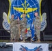 Three military personnel in camouflage uniforms are participating in what appears to be a change of command or guidon ceremony on a stage. A large unit emblem banner is displayed in the background featuring a globe, wings, and lightning bolts consistent with Air Mobility Command insignia. A banner reading 'Fort Dawg Sister Brotherhood' is visible at the front of the stage.
