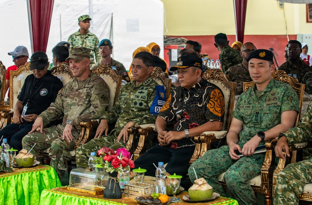 A multinational military gathering showing officers and officials from multiple nations seated in ornate golden chairs under a tent canopy. Visible name tags and uniform patches identify representatives from the U.S. Army, TNI AD (Indonesian Army), and Singapore Armed Forces, suggesting a joint military exercise or ceremony. The formal seating arrangement and decorative table settings indicate an official diplomatic or ceremonial event.