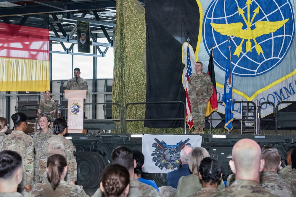 A military ceremony is taking place inside what appears to be an aircraft hangar, likely at a US Air Force base in Germany based on the visible German and US flags. A uniformed service member is speaking at a podium while another stands at attention on stage, with a large audience of military personnel in camouflage uniforms watching. The backdrop includes a large US Air Force command insignia banner and camouflage netting.