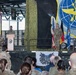 A military ceremony is taking place inside what appears to be an aircraft hangar, likely at a US Air Force base in Germany based on the visible German and US flags. A uniformed service member is speaking at a podium while another stands at attention on stage, with a large audience of military personnel in camouflage uniforms watching. The backdrop includes a large US Air Force command insignia banner and camouflage netting.