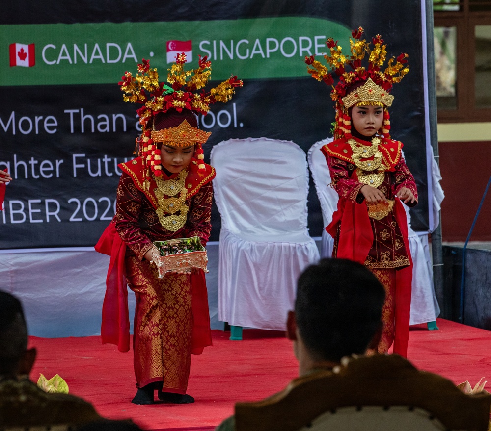 Two young children are performing a traditional cultural dance on a red-carpeted stage, dressed in elaborate red and gold traditional costumes with ornate headdresses. The performance appears to be part of a formal event involving Canada and Singapore, as indicated by the banner visible in the background. The event appears to be school-related, with text referencing 'More Than a School' and 'Brighter Future' with a date of 2020.