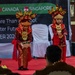 Two young children are performing a traditional cultural dance on a red-carpeted stage, dressed in elaborate red and gold traditional costumes with ornate headdresses. The performance appears to be part of a formal event involving Canada and Singapore, as indicated by the banner visible in the background. The event appears to be school-related, with text referencing 'More Than a School' and 'Brighter Future' with a date of 2020.