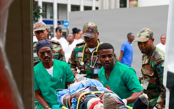 A medical emergency scene showing a patient being transported on a stretcher toward what appears to be an ambulance or medical vehicle. Multiple personnel in green medical scrubs and military camouflage uniforms are working together to move the patient. The setting appears to be outside a medical facility.