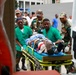 A medical emergency scene showing a patient being transported on a stretcher toward what appears to be an ambulance or medical vehicle. Multiple personnel in green medical scrubs and military camouflage uniforms are working together to move the patient. The setting appears to be outside a medical facility.
