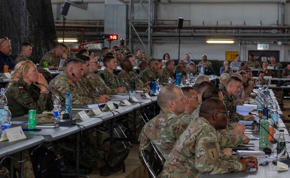 A large military briefing or conference is taking place inside what appears to be a hangar or large industrial facility. Numerous military personnel in camouflage uniforms, representing multiple nations, are seated at long tables facing a presenter or screen. The setting suggests a multinational military planning or coordination event.