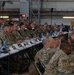 A large military briefing or conference is taking place inside what appears to be a hangar or large industrial facility. Numerous military personnel in camouflage uniforms, representing multiple nations, are seated at long tables facing a presenter or screen. The setting suggests a multinational military planning or coordination event.