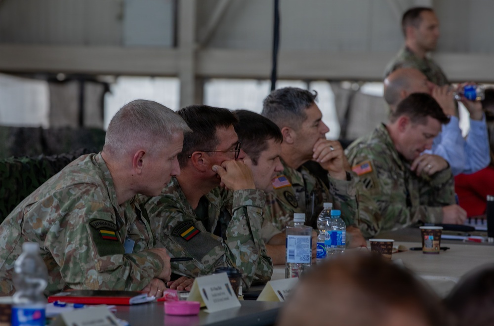 A group of military personnel from multiple nations are seated at a table during what appears to be a briefing or conference. At least two soldiers are wearing Lithuanian flag patches on their camouflage uniforms, while others display US Army patches. The setting appears to be a military tent or hangar facility.