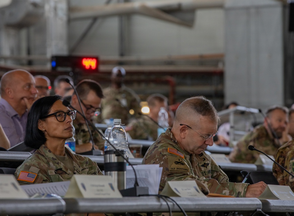 A military briefing or conference is taking place in what appears to be a large hangar or industrial facility. Multiple U.S. Army personnel in OCP (Operational Camouflage Pattern) uniforms are seated at tables with documents, microphones, and water bottles. The foreground features two soldiers prominently, one with visible 'Old Ironsides' (1st Armored Division) unit patch.