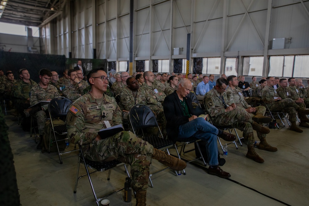 A large group of U.S. Army soldiers in OCP (Operational Camouflage Pattern) uniforms are seated in rows of folding chairs inside what appears to be a military hangar or large facility. The audience appears to be attending a briefing, ceremony, or formal presentation. A small number of civilians are also present among the predominantly uniformed attendees.