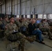 A large group of U.S. Army soldiers in OCP (Operational Camouflage Pattern) uniforms are seated in rows of folding chairs inside what appears to be a military hangar or large facility. The audience appears to be attending a briefing, ceremony, or formal presentation. A small number of civilians are also present among the predominantly uniformed attendees.