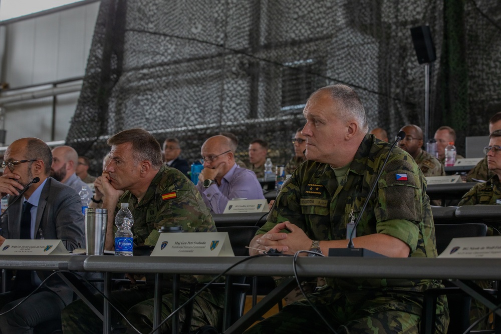 A multinational military conference or briefing is taking place in what appears to be a military hangar or field facility decorated with camouflage netting. Several senior military officers from different nations, including Spain and the Czech Republic, are seated at tables alongside civilian officials. Name placards are visible on the tables, identifying at least one participant as Maj Gen Petr Svoboda, Territorial Forces Command Commander.